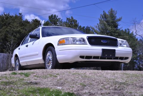 2002 Ford Crown Victoria - A white 2002 Ford Crown Victoria is viewed from a low angle on a grassy hill under a partly cloudy sky.