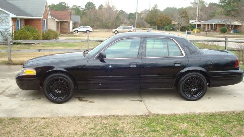 2004 Ford Crown Victoria - A black sedan with custom black rims sits parked on a concrete driveway in front of a residential house.