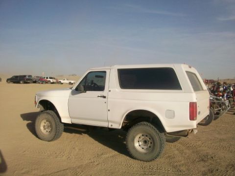 1996 Ford Bronco - A white SUV is parked in a sandy area with other vehicles in the background under a clear blue sky.