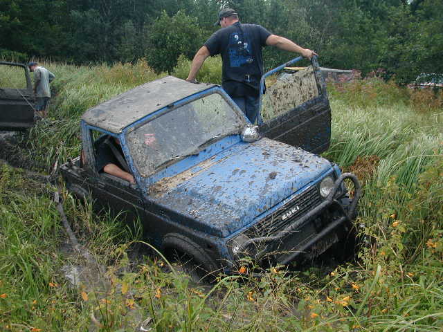 Blue Suzuki Samurai with ZUKI hood lettering sunk in a grassy mud bog, door open
