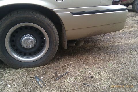 2000 Mercury Grand Marquis - The rear wheel and exhaust pipe of a gold Mercury Grand Marquis are visible, with another vehicle in the background.