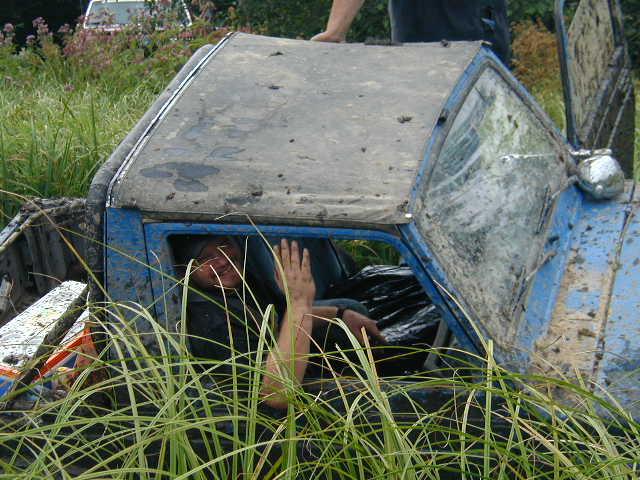 Muddy blue pickup truck stuck in grass, person's head visible through window. Another vehicle in background