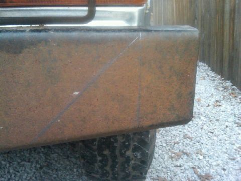 1987 Ford Bronco - A close-up view of a heavily rusted bumper and a tire, with a wooden fence in the background.