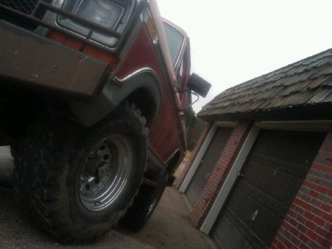 1987 Ford Bronco - This red SUV features large tires and an aftermarket bumper, seen parked beside a brick garage.