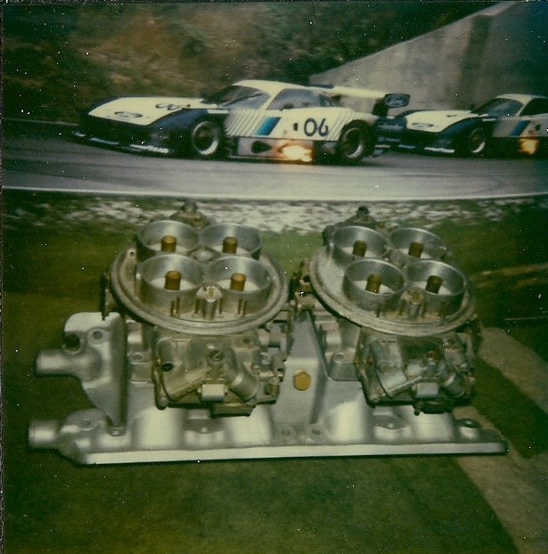 Dual carburetors mounted on an aluminum intake manifold, with race cars in the background