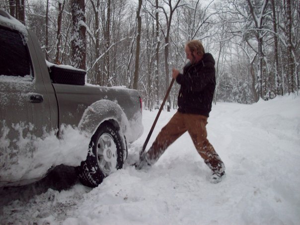 Person shoveling snow around the rear wheel of a stuck pickup truck in a wooded area