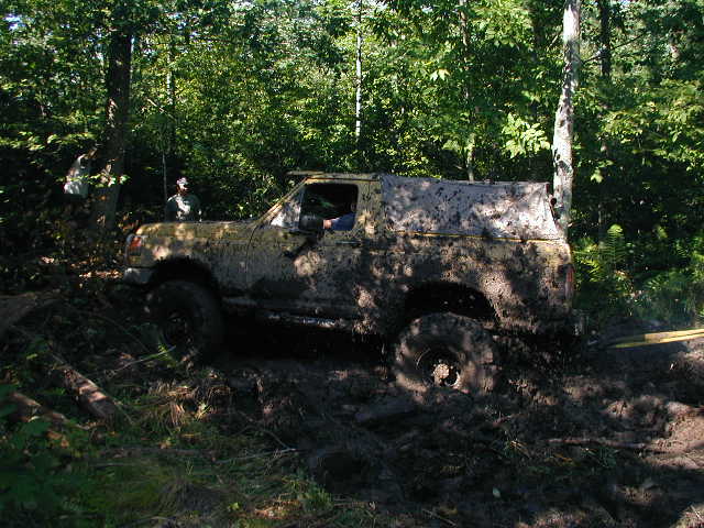 Mud-covered yellow SUV stuck in mud with trees in background. Visible text: SUPERMOTORS