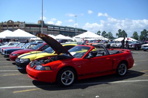 2002 Ford Mustang - A line of cars, including a red Mustang GT convertible, are parked outside under a sunny sky with tents and a stadium in the background.