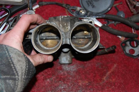 1994 Ford Bronco - A hand holds a dual throttle body, with tools and other engine parts scattered in the background.