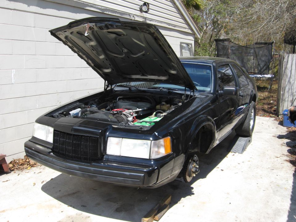 Black Lincoln Mark VII coupe with hood open and front wheels off in a driveway