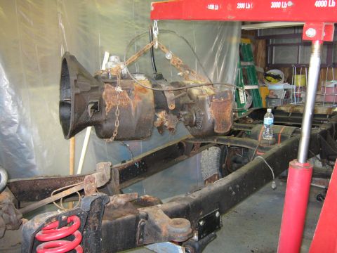 1977 Ford F-150 - A heavily rusted transmission is suspended by chains from an engine hoist over the frame of a truck.