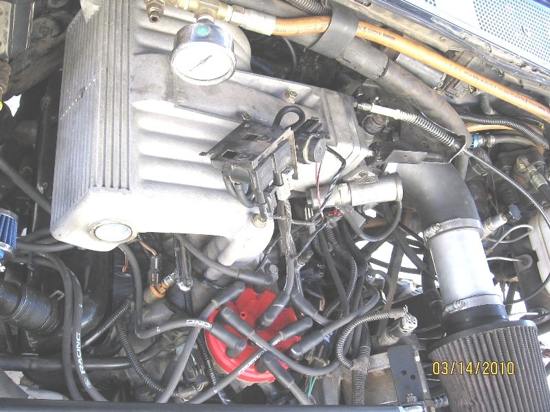 Engine bay with valve cover, mounted gauge, red distributor cap, black intake tubing and blue cone filter