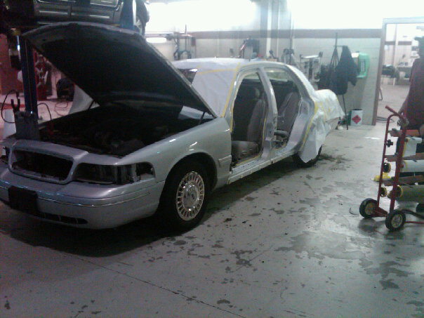 White Crown Victoria in body shop with hood up, masked with yellow tape for paint prep
