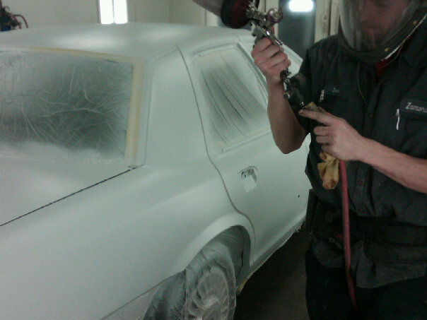 Worker in respirator spray-painting primer on driver's side of a masked sedan in a paint booth