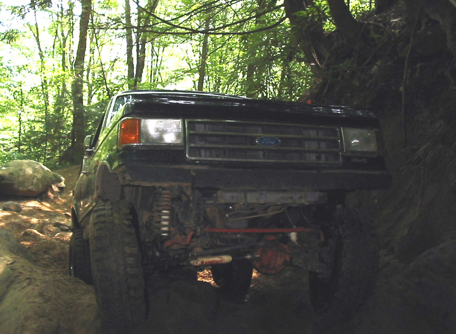 Dark blue lifted Ford Bronco front view off-roading on rocks in forest, showing axle and Rancho stabilizer