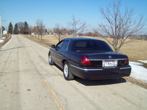 1999 Mercury Grand Marquis - A dark sedan is parked on the side of a country road with bare trees and a clear blue sky.
