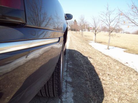1999 Mercury Grand Marquis - Side profile of a 1999 Mercury Grand Marquis on a dirt road with trees and snow patches.