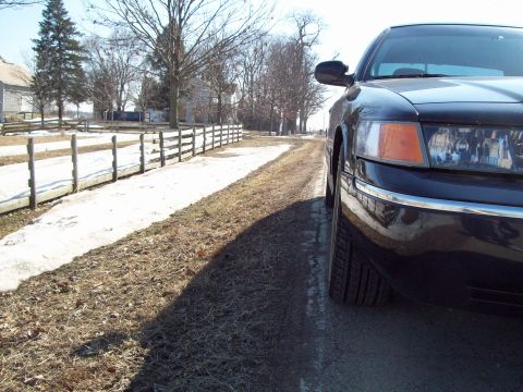1999 Mercury Grand Marquis - A black sedan is parked on the side of a rural road with snow on the ground and a wooden fence.
