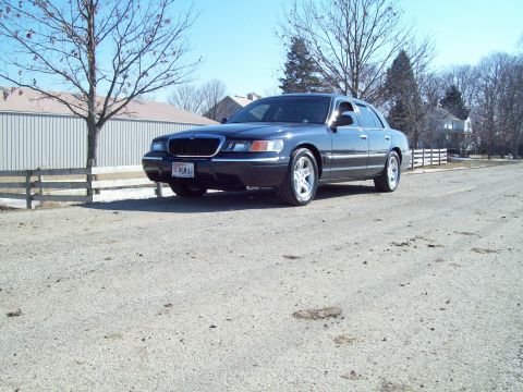 1999 Mercury Grand Marquis - A Mercury Grand Marquis is parked on a gravel road with a building and trees in the background.