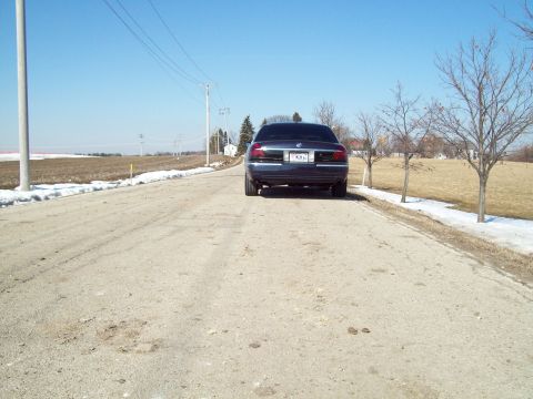1999 Mercury Grand Marquis - Rear view of a dark sedan on a rural dirt road under a clear blue sky.