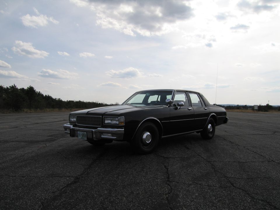Black sedan parked on cracked asphalt lot, cloudy sky overhead. Appears to be a late 1970s or early 1980s model