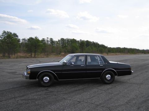 1991 Ford LTD Crown Victoria - A black sedan with chrome accents and steel wheels is parked on an asphalt surface with trees in the background.