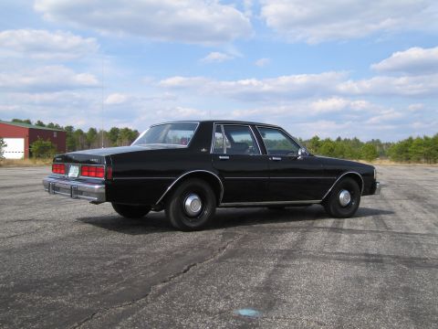 1991 Ford LTD Crown Victoria - This black sedan is parked outdoors on a cracked asphalt surface under a partly cloudy sky.