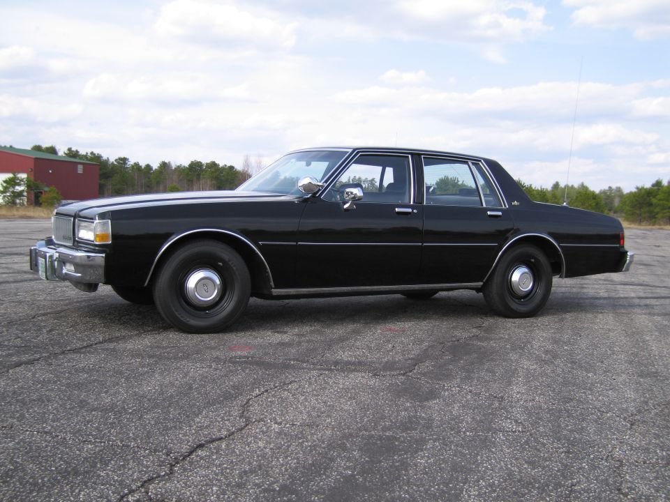 Black sedan parked on asphalt lot, side view. Appears to be a 1980s model