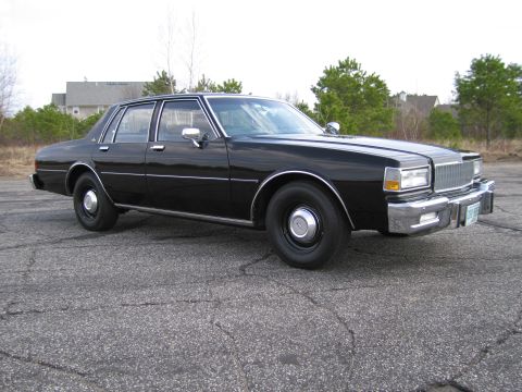 1991 Ford LTD Crown Victoria - This black sedan is parked on a cracked asphalt surface with trees and houses visible in the background.