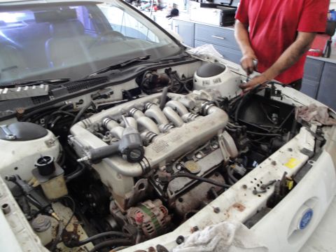 1989 Ford Taurus - The engine bay of a white Ford Taurus is shown with a SHO engine and an impact wrench. A person is working on the engine.