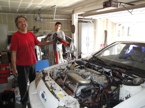 1989 Ford Taurus - Two men work on the engine bay of a 1989 Ford Taurus in a garage, with the engine partially removed.