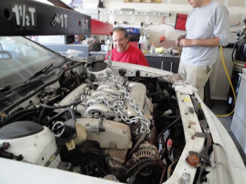 1989 Ford Taurus - Mechanics use an engine hoist to remove the engine from a white car in a garage.