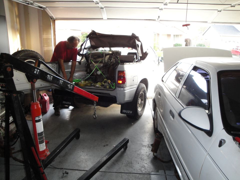 Man in red shirt uses engine hoist to remove engine from silver Isuzu pickup truck bed. White sedan parked next to it