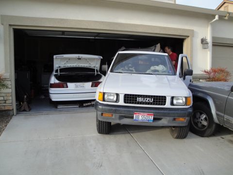 1989 Ford Taurus - Two vehicles are parked in a driveway, one a white Isuzu pickup and the other a white car with its trunk open.
