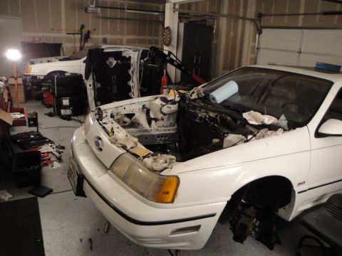 1989 Ford Taurus - A white 1989 Ford Taurus sits in a garage with its hood open, revealing an exposed engine bay and undergoing maintenance.