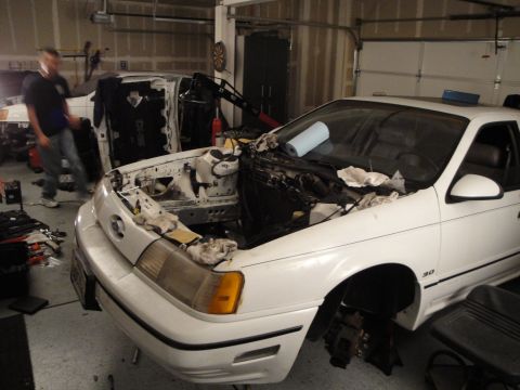 1989 Ford Taurus - A white sedan is in a garage with its engine removed, and another vehicle is partially visible in the background.