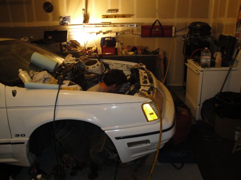 1989 Ford Taurus - A person works on the engine of a white car in a garage setting.