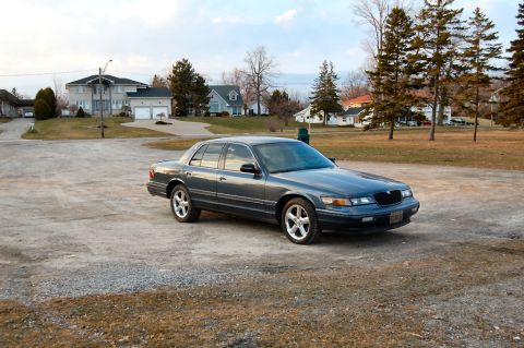 1985 Mercury Grand Marquis - This dark gray sedan, likely a 1995 Mercury Grand Marquis, is parked on a gravel lot with houses in the background.