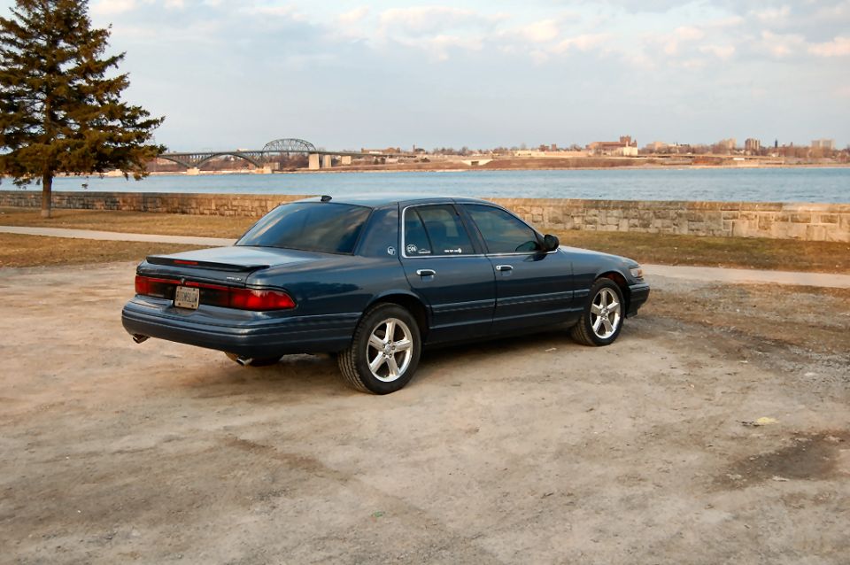 Dark blue sedan parked on dirt lot near water, bridge in background