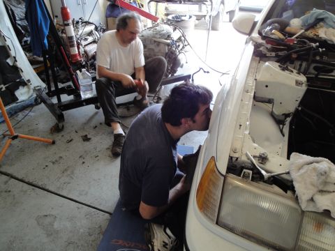 1989 Ford Taurus - Two mechanics are working on the engine of a white car in a cluttered garage.