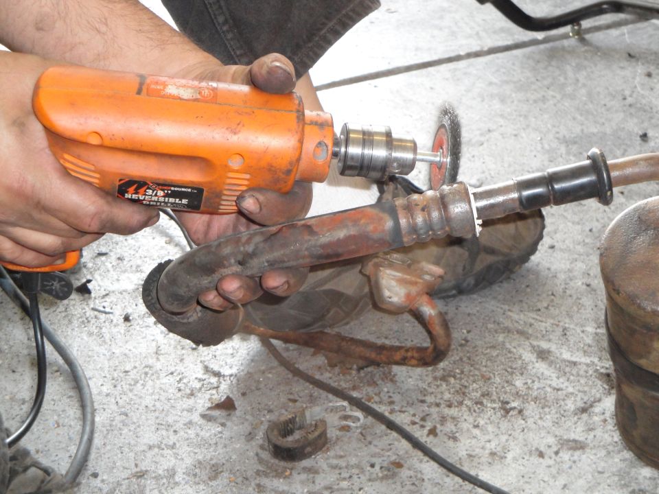 Hands holding orange drill with wire brush attachment cleaning a rusty, bent metal pipe