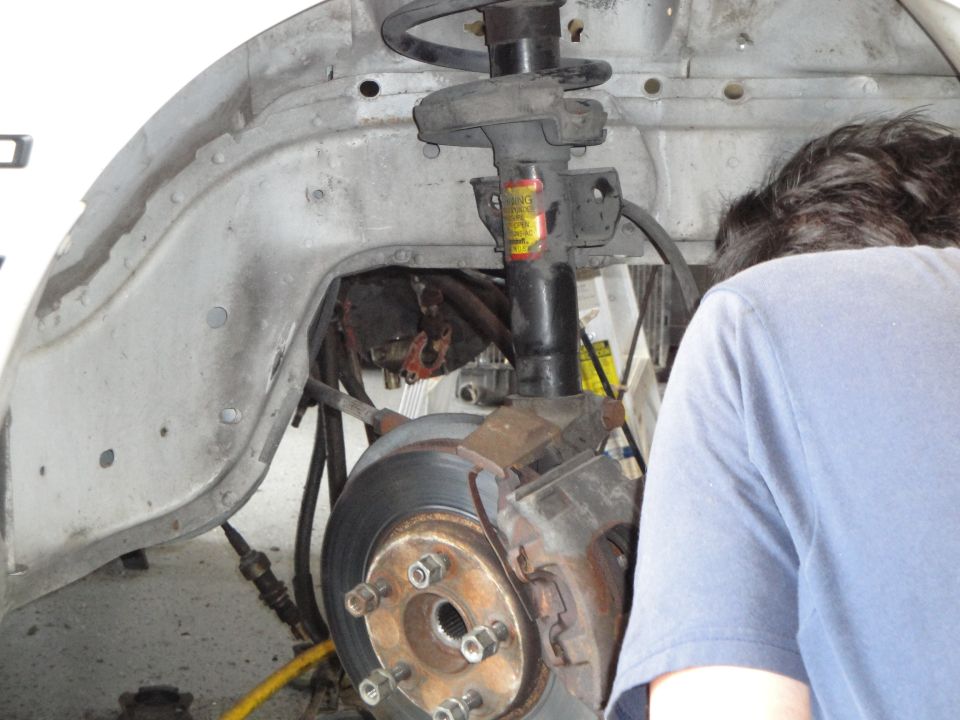 Car's front wheel well and suspension system with a person working on it. Rusty brake rotor and caliper visible