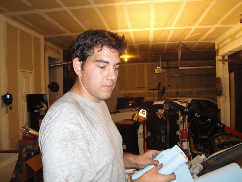 Man holding paper towels in a cluttered garage with an engine hoist and car parts visible