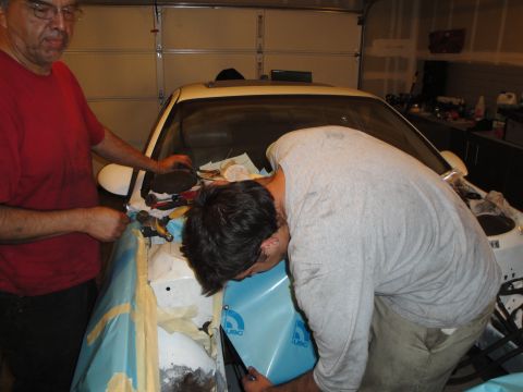 1989 Ford Taurus - Two individuals are seen working on the front fender area of a white vehicle, with blue protective sheeting applied.