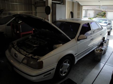 1989 Ford Taurus - A white 1989 Ford Taurus is parked in a garage with its hood open, revealing the engine compartment. Another vehicle is visible in the back…