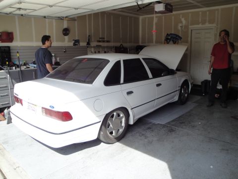 1989 Ford Taurus - A white 1989 Ford Taurus sedan is parked in a garage with its hood open, while two people stand nearby.