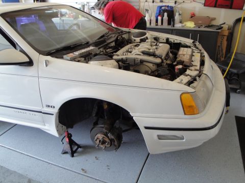 1989 Ford Taurus - A person works on the engine of a white sedan that is elevated on jack stands in a garage.