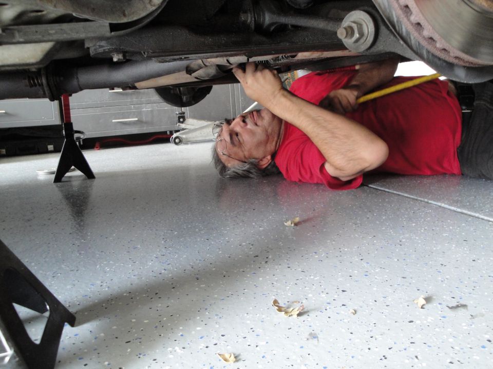 Guy in red shirt lying on garage floor, working on the underside of a car supported by jack stands. He's holding a yellow tool