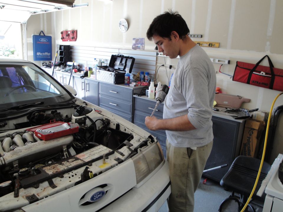 White Ford sedan with hood open, showing engine and a man with a drill. Garage setting