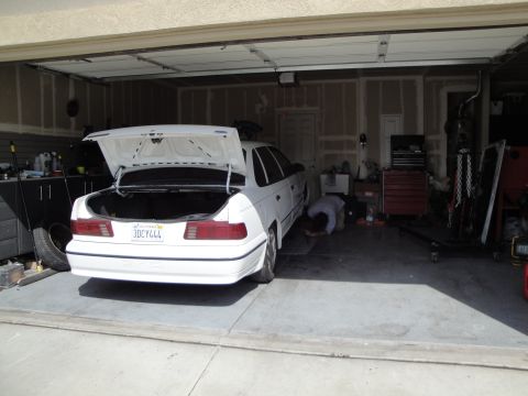 1989 Ford Taurus - A white 1989 Ford Taurus is parked in a garage with its trunk open while someone works underneath it.